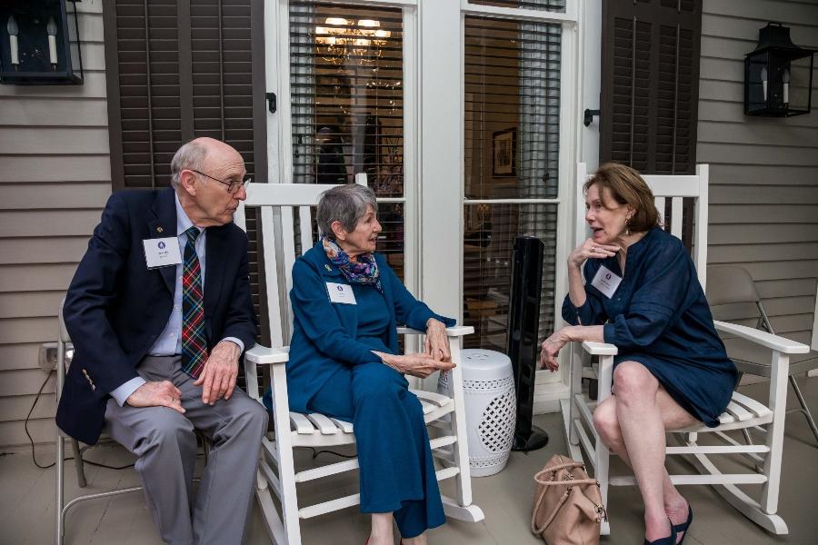 Three people sit on porch rocking chairs while talking. 